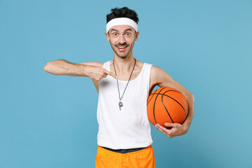 Excited fun young man basketball player with thin skinny body sportsman in headband shirt shorts whistle hold basketball ball posing isolated on blue background. Workout gym sport motivation concept