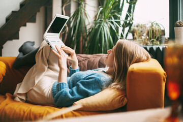 A young girl is lying on the sofa, holding a laptop on her lap and texting with friends and colleagues. The girl works at home at the computer. Work online, freelance, social distance
