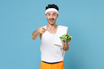 Excited sporty man with thin skinny body sportsman in headband shirt shorts hold vegetable salad in bowl point index finger on camera isolated on blue background. Workout gym sport motivation concept.
