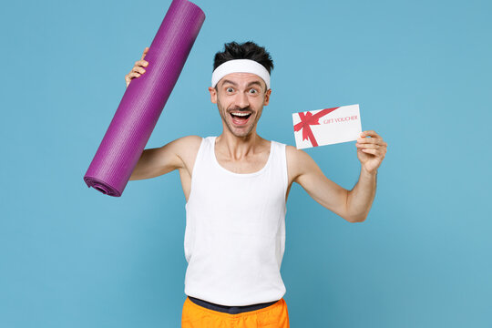 Surprised Young Strong Sporty Fitness Man With Skinny Body Sportsman In White Headband Shirt Shorts Hold Yoga Mat Gift Certificate Isolated On Blue Background. Workout Gym Sport Motivation Concept.