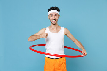 Excited young sporty fitness man with thin skinny body sportsman in headband shirt shorts training with hula hoop showing thumb up isolated on blue background. Workout gym sport motivation concept.