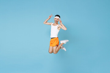 Obraz premium Full length portrait of cheerful young fitness man with skinny body sportsman in headband shirt shorts jumping showing victory sign isolated on blue background. Workout gym sport motivation concept.