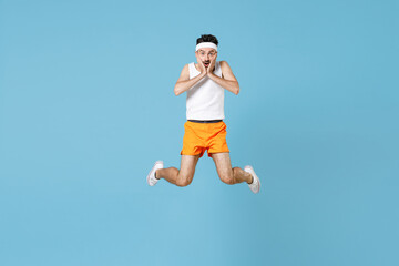 Fototapeta na wymiar Full length portrait of shocked young fitness man with skinny body sportsman in headband shirt shorts jumping put hands on cheeks isolated on blue background. Workout gym sport motivation concept.
