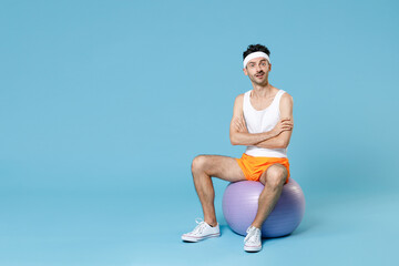 Full length portrait of amazed young man with skinny body sportsman in headband shirt shorts sit on fitball holding hands crossed isolated on blue background. Workout gym sport motivation concept.