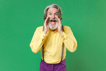 Excited cheerful elderly gray-haired mustache bearded man in casual yellow shirt suspenders screaming with hands gesture near mouth looking camera isolated on green colour background, studio portrait.