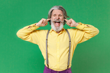 Crazy elderly gray-haired mustache bearded man in casual yellow shirt suspenders covering ears with fingers keeping eyes closed screaming isolated on bright green colour background, studio portrait.