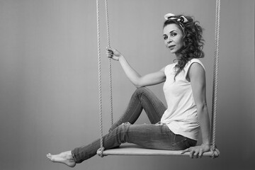 Beautiful brunette girl with long curly hair with a bow barefoot in ripped jeans and a white T-shirt sits on a swing in the studio
