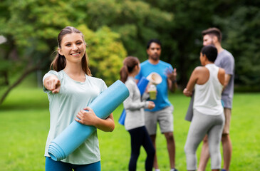 fitness, sport and healthy lifestyle concept - happy smiling young woman with mat pointing finger to camera over group of people meeting for yoga class at summer park