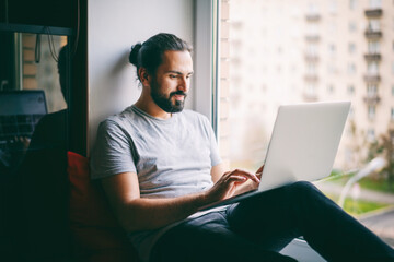 Handsome attractive young dark-haired man working on laptop at home while sitting on windowsill...