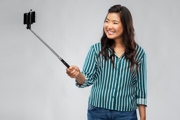 technology and people concept - happy asian woman taking picture with smartphone on selfie stick over grey background