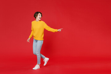 Fototapeta na wymiar Full length side view of excited funny young brunette woman 20s wearing basic yellow sweater pointing index finger aside on mock up copy space isolated on bright red colour background studio portrait.