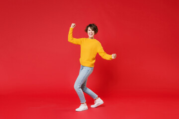 Full length side view of happy joyful laughing young brunette woman 20s wearing casual yellow sweater clenching fists doing winner gesture isolated on bright red colour background studio portrait.