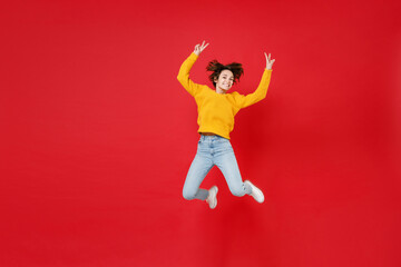 Full length of smiling cheerful funny young brunette woman 20s wearing basic casual yellow sweater jumping rising hands showing victory sign isolated on bright red colour background studio portrait.