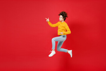 Full length side view of cheerful young brunette woman 20s in basic yellow sweater jumping pointing index fingers aside on mock up copy space isolated on bright red colour background studio portrait.