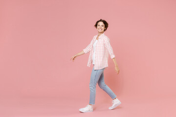 Fototapeta na wymiar Full length side view of smiling beautiful attractive young woman 20s wearing casual basic checkered shirt walking going looking camera isolated on pastel pink colour background, studio portrait.