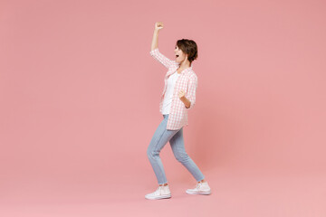 Fototapeta na wymiar Full length side view of excited happy young brunette woman 20s wearing casual checkered shirt standing clenching fists doing winner gesture isolated on pastel pink colour background, studio portrait.