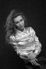 A beautiful slender brunette with long curly hair in a white satin blouse sits in the studio on a black background, bw