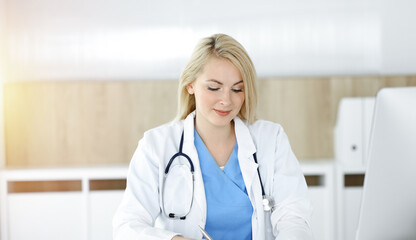 Woman-doctor at work while sitting at the desk in sunny clinic. Blonde cheerful physician filling up medication history record form