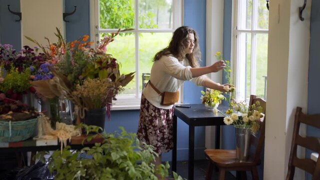 Woman florist with long loose hair takes picture of small bouquet and works with fresh yellow flowers at table near window at floral masterclass