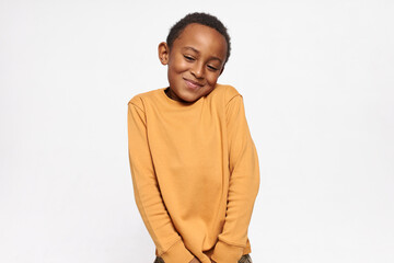 Horizontal shot of cute handsome dark skinned little boy shrugging shoulders feeling embarrassed with uncomfortable question, looking down with shy timid smile. Human emotions and body language