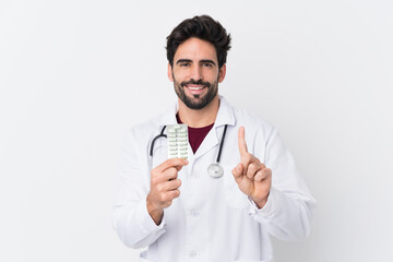 Young handsome man with beard over isolated white background wearing a doctor gown and holding pills