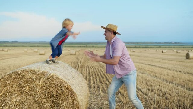 happy childhood, little joyful male child enjoys playing with his father jumping from haystack outdoors in countryside