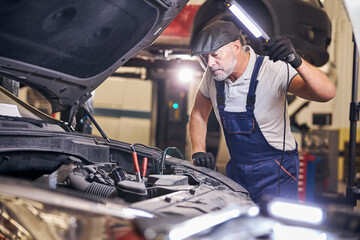 Bearded auto mechanic inspecting car engine at service station