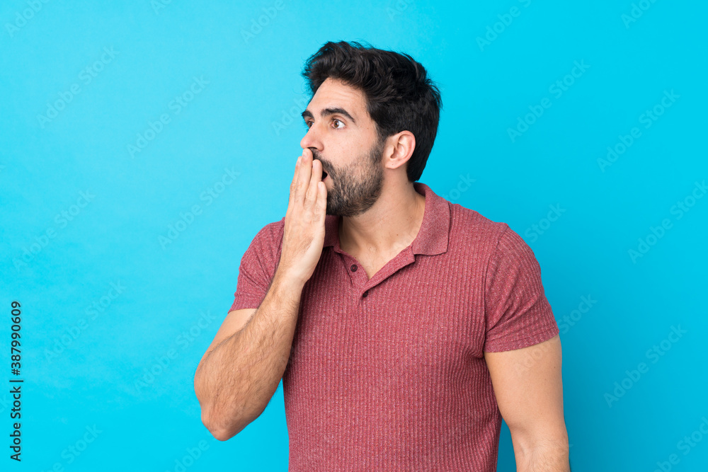 Wall mural Young handsome man with beard over isolated blue background yawning and covering wide open mouth with hand