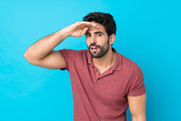 Young handsome man with beard over isolated blue background looking far away with hand to look something