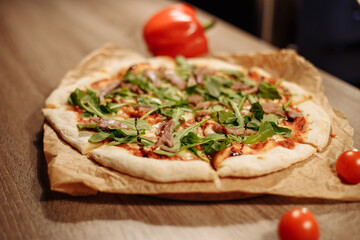 fresh pizza with herbs, tuna and balsamic sauce on food paper on a wooden table in a cozy cafe, bell peppers in the background and cherry tomatoes in the foreground