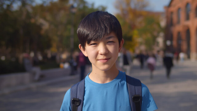 Asian schoolboy carrying backpack and smiling at camera while posing outdoors