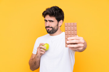 Young handsome man with beard over isolated yellow background taking a chocolate tablet in one hand and an apple in the other