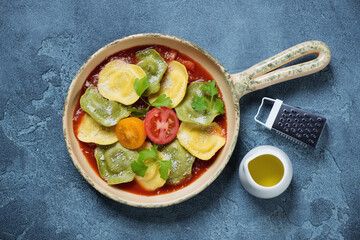 Beige serving pan with ravioli on tomato sauce bedding, flatlay over blue stone background