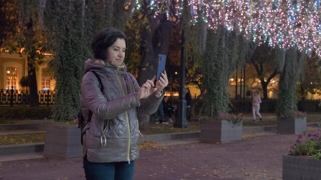 A middle-aged woman in a lilac jacket photographs street decorations on a smartphone. Vokrut New Year's illumination. in evening time