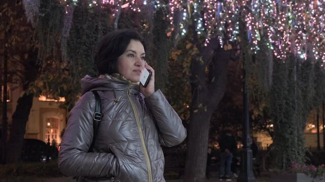 A middle-aged Caucasian woman in a lilac jacket takes out a smartphone and talks. New Year's illumination. in evening time