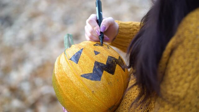 Close up woman hands paint orange pumpkin with black paint in autumn forest.