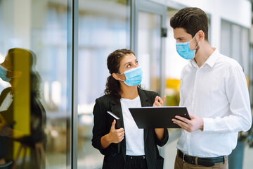 Two colleagues in protective face masks in modern office discussing together work issues. Business People back at work in office after quarantine. Covid-19.