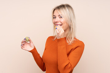 Young blonde Russian woman over isolated background holding colorful French macarons and eating it