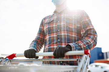 Young man stands outside the supermarket with the trolley wearing sterile medical mask and gloves to protect from coronavirus. Customer at a shops parking is going for purchases. Healthcare concept.