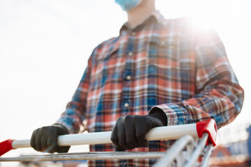 Young man stands outside the supermarket with the trolley wearing sterile medical mask and gloves to protect from coronavirus. Customer at a shops parking is going for purchases. Healthcare concept.