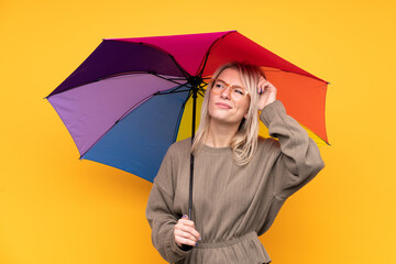 Young blonde woman holding an umbrella over isolated yellow wall having doubts and with confuse...