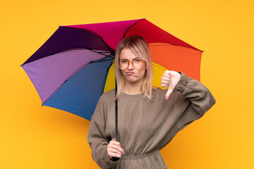 Young blonde woman holding an umbrella over isolated yellow wall showing thumb down sign