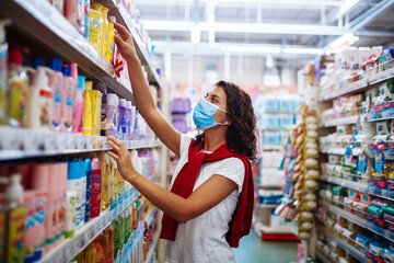 Young woman picks up the disinfecting and cleaning scour at a supermarket wearing a medical sterile mask during the coronavirus pandemic quarantine. Healthcare and home sanitizing concept.