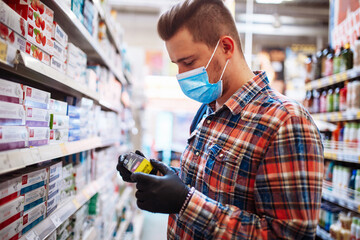 Young man is choosing a toothpaste at a supermarket wearing a medical sterile mask during the coronavirus pandemic quarantine. Healthcare, hygiene and home sanitizing concept.