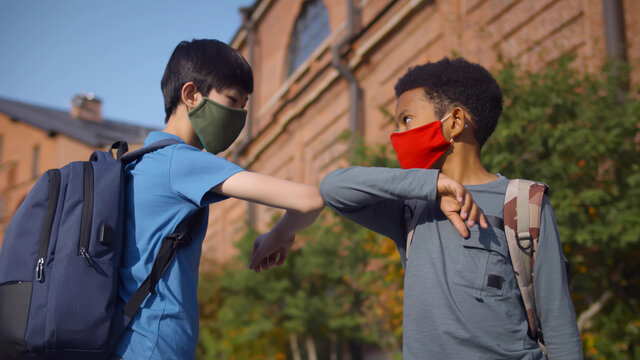 Side view of diverse schoolboys in protective mask doing elbow greeting outdoors