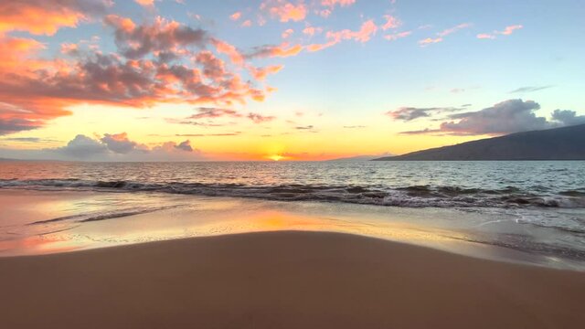 Wide shot of gentle waves rolling to shore in a tropical island sunset with sandy foreground and a distant island in the distance with fluffy pink clouds set against a light blue sky