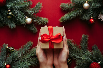 Craft gift with a red ribbon in the hands of a young girl with a nude manicure next to the branches of a Christmas tree. The concept of giving and receiving gifts. Christmas and New Year