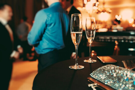 Official Corporate Banquet Dinner. Two Glasses With Champagne, Shiny Womens Handbag And Phone On The Table. Company Employees On The Background