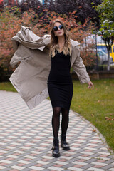 attractive slim young brunette in fashionable trench, black dress and sunglasses in park. autumn, yellow leaves.