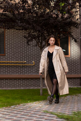 attractive slim young brunette in fashionable trench coat and black dress walking towards. autumn, yellow leaves. brick building background.
